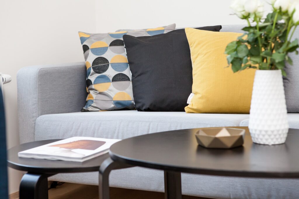 Un sofá con varios almohadones detrás de dos mesas con una revista, un cuenco y un jarrón de flores